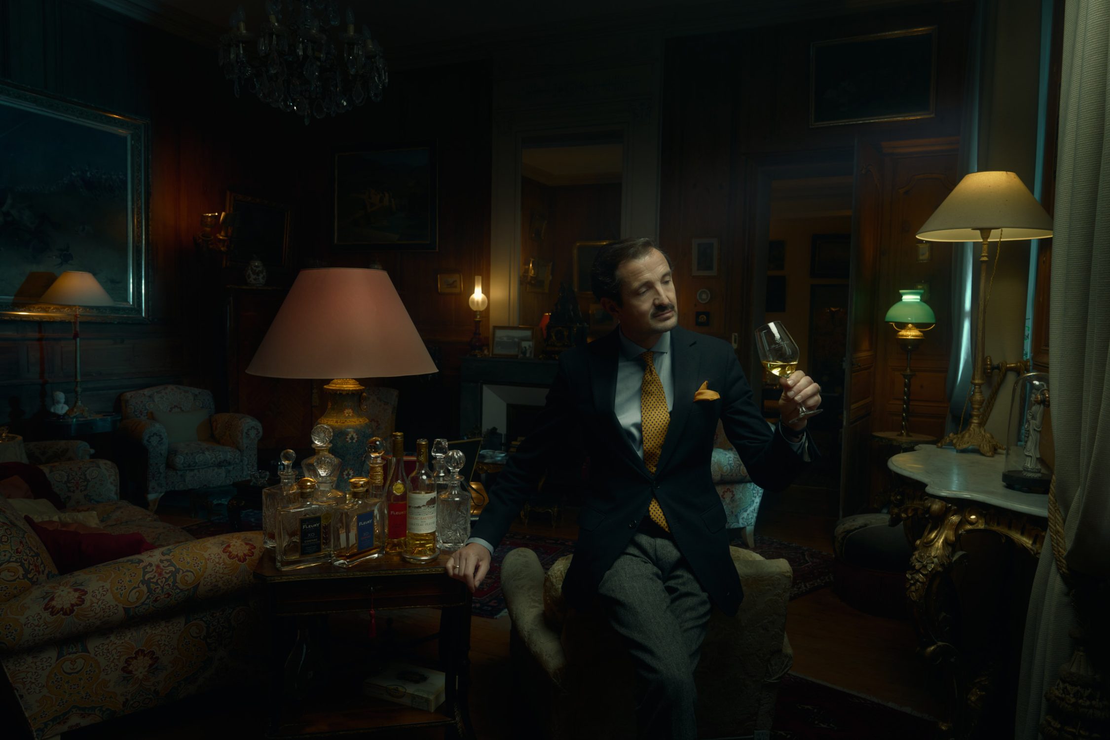 Olivier Fleury seated with a glass in a richly furnished room lit by table lamps.